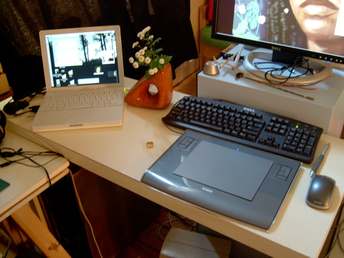 A work desk: white iBook, black keyboard, Wacom tablet and mouse, a Dell monitor showing a portrait in progress, daisies in an orange vase