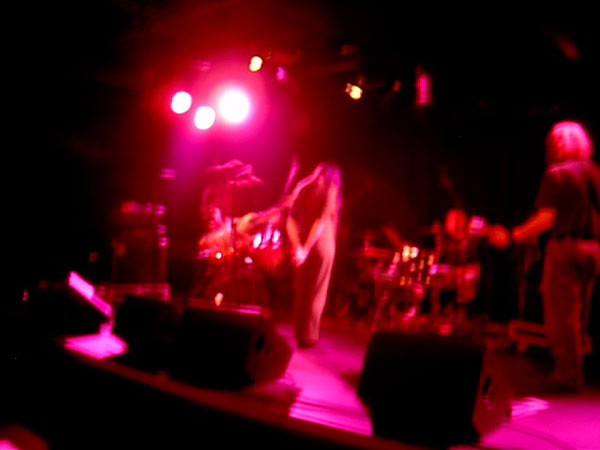 Jarboe on stage in Rotterdam, blurred in magenta light, long-haired guitarist at right and drum kit behind