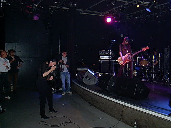 Concert at a small venue: singer bent over at the stage edge in black, guitarist and drummer on stage, sparse audience watching