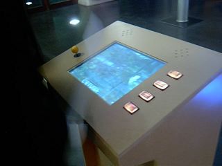 The Endless Forest running on a museum kiosk: metal arcade cabinet with yellow joystick, four buttons and a recessed screen of forest