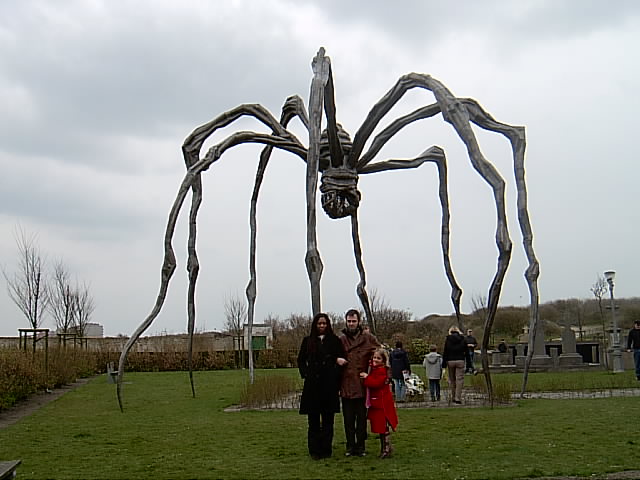 Family posing under Louise Bourgeois's giant spider Maman: a woman in black, a man, and a small girl in a red coat, grey sky above