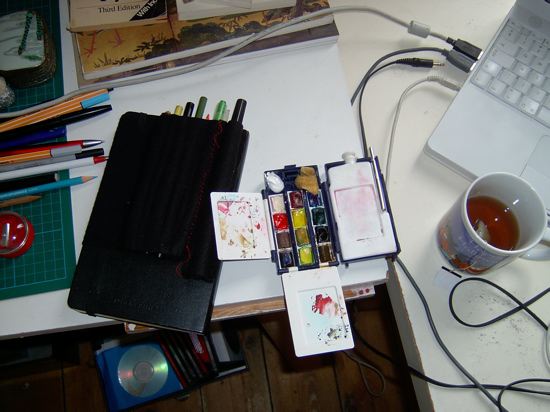 Portable studio on a desk: black felt pencil roll, tiny travel watercolor set with stained mixing lids, sketchbook, tea mug and laptop