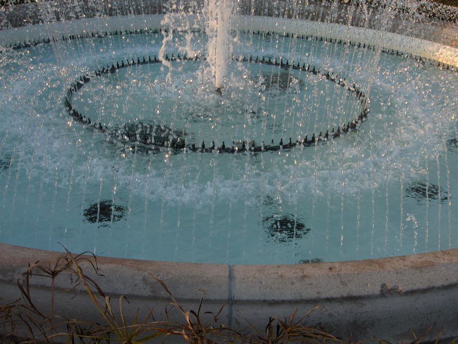 Circular fountain basin ringed by thin jets of water catching evening light, dry grasses in the foreground