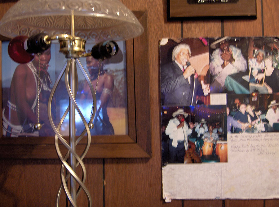 Wood-panelled wall of framed personal photos — a man in a white suit singing, band snapshots with congas — behind a frosted-glass lamp