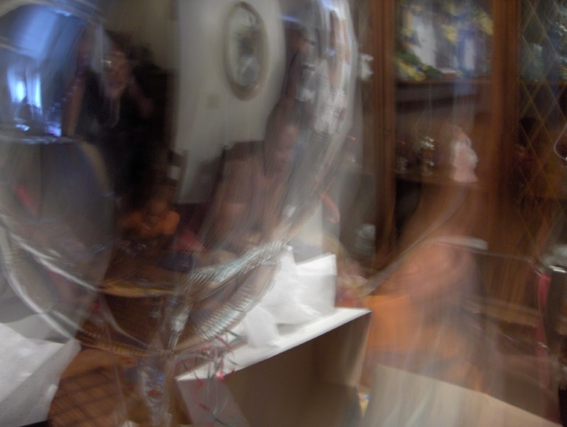 Long-exposure blur of a family living room seen through a mylar balloon — clock, cabinet and figures smeared into ghosts