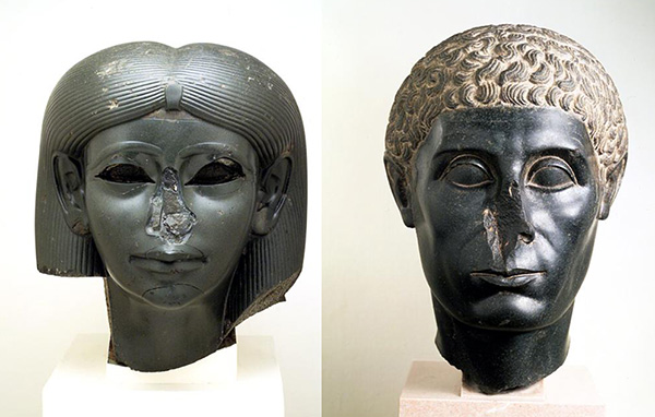 Two ancient Egyptian black stone heads with damaged noses, side by side on museum stands — one with wig, one curly-haired