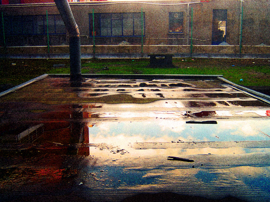 Rain water pooled on a flat surface reflecting the sky, a drainpipe and grass behind, an old school building beyond the fence