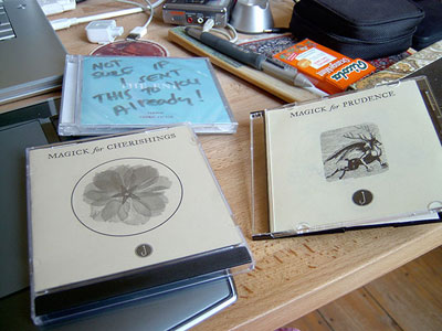 Three Jandek-style CD-Rs on a desk: 'Magick for Cherishings' and 'Magick for Prudence' with pressed-flower covers, one disc hand-labeled 'NOT SURE IF I SENT THIS TO YOU ALREADY!'
