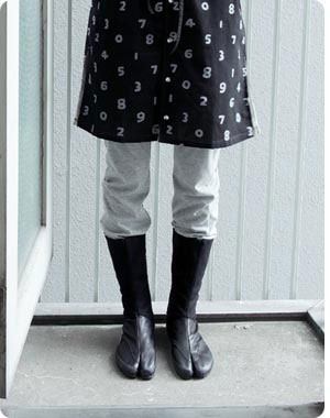 Standing in split-toe tabi boots on a concrete doorstep, grey trousers pushed into the black boots, a number-printed black skirt above Standing in split-toe tabi boots on a concrete doorstep, grey trousers pushed into the black boots, a number-printed black skirt above