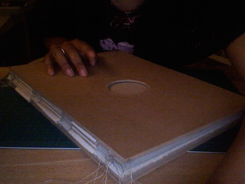 Bookbinding in progress: a hand steadies a cardboard book cover with a circular window cut out, exposed cords at the raw spine
