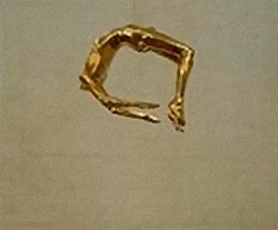 Grainy still of a small gold figure curled into a ring, floating mid-air against beige — falling gilded body