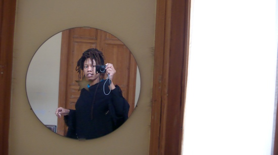 Self-portrait in a round mirror by the wooden door, camera at eye, in a black sweater with dreadlocks tied back