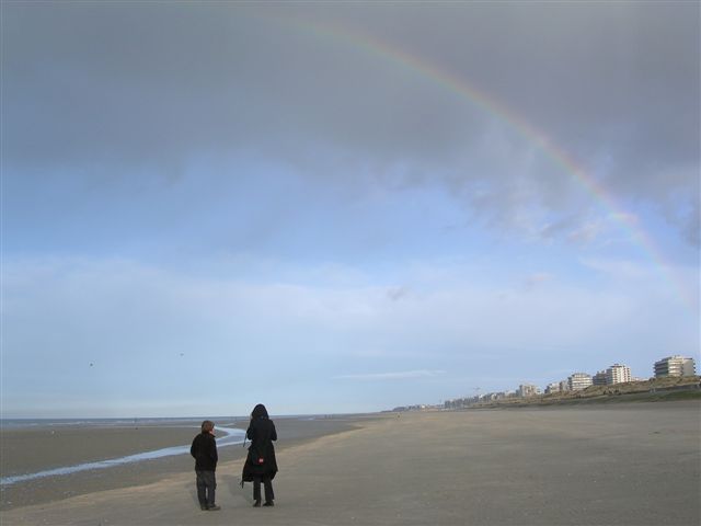 Two figures on the wide wet sand of the Belgian coast under a heavy sky, a rainbow arcing down toward distant apartment blocks