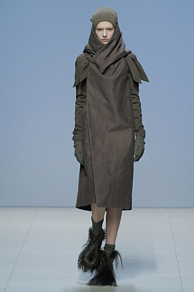 Rick Owens–style runway look: model wrapped in a grey-brown scarf-dress with knit cap, long gloves, and shaggy fur boots