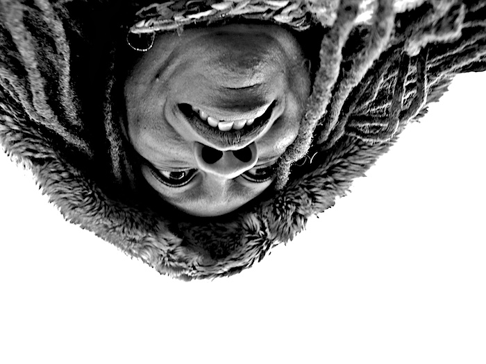 Upside-down black-and-white portrait: a smiling face hanging inverted out of a hood of dreadlocks against white