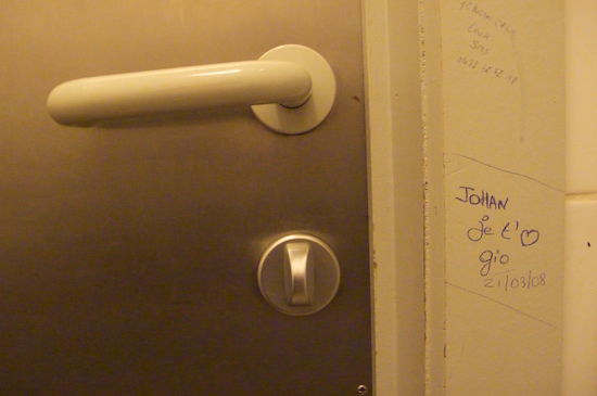Hotel-room door detail: steel panel with white handle, ballpoint graffiti on the frame — 'JOHAN Je t'♥ gio 21/03/08'