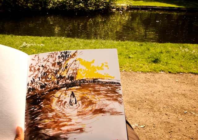 Sketchbook held up in a park: brown-ink drawing of the pond with a dark figure's ripples, the real pond and lawn behind it