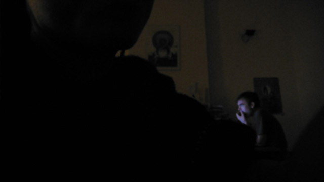 Almost-black room: a dark silhouetted head in the foreground, a man's face lit by a screen at the far desk, an icon poster on the wall