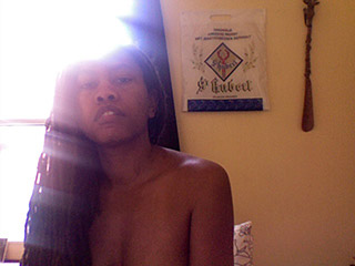Washed-out webcam self-portrait, bare-shouldered with a light flare, a St Hubert pilgrimage banner on the wall behind