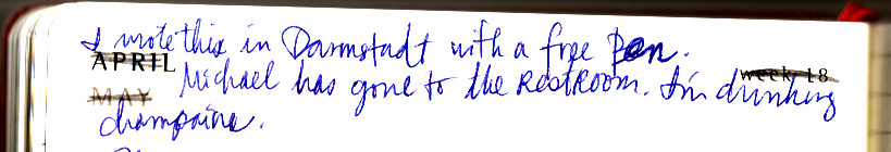 Diary line in blue ballpoint over the printed word APRIL: 'I wrote this in Darmstadt with a free pen. Michael has gone to the restroom. I'm drinking champagne.'