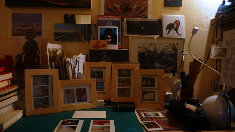Studio corner crowded with wood frames holding small game-screenshot prints, art postcards pinned above a green cutting mat