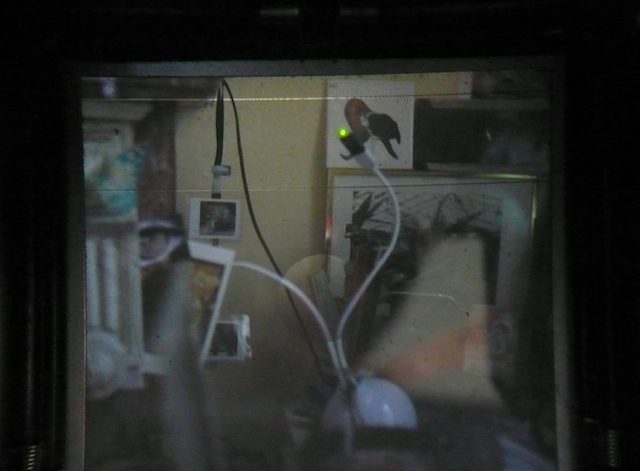 Camera-viewfinder framing of a studio wall: a webcam and USB lamp plugged in, small prints taped up, everything dim and greenish Camera-viewfinder framing of a studio wall: a webcam and USB lamp plugged in, small prints taped up, everything dim and greenish