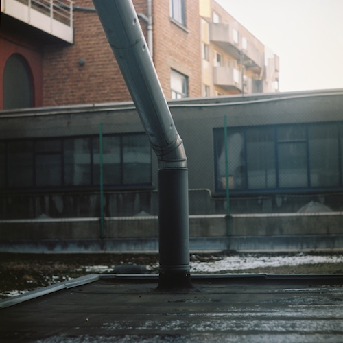 Square medium-format photo: a drainpipe kinking down onto a flat tar roof, brick school building and fenced windows behind, morning haze Square medium-format photo: a drainpipe kinking down onto a flat tar roof, brick school building and fenced windows behind, morning haze