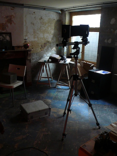 Bare studio with peeling walls: a large-format camera on a tripod facing a trestle table, cold window light, paint-spattered floor Bare studio with peeling walls: a large-format camera on a tripod facing a trestle table, cold window light, paint-spattered floor