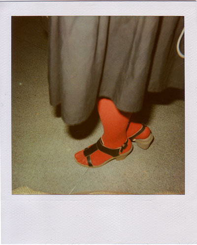 Polaroid of a foot in a bright red-orange stocking and black strappy sandal, dark skirt hem swinging above speckled lino Polaroid of a foot in a bright red-orange stocking and black strappy sandal, dark skirt hem swinging above speckled lino