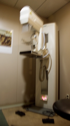Blurred photo of a mammogram machine in a beige clinic room, footswitches on the floor