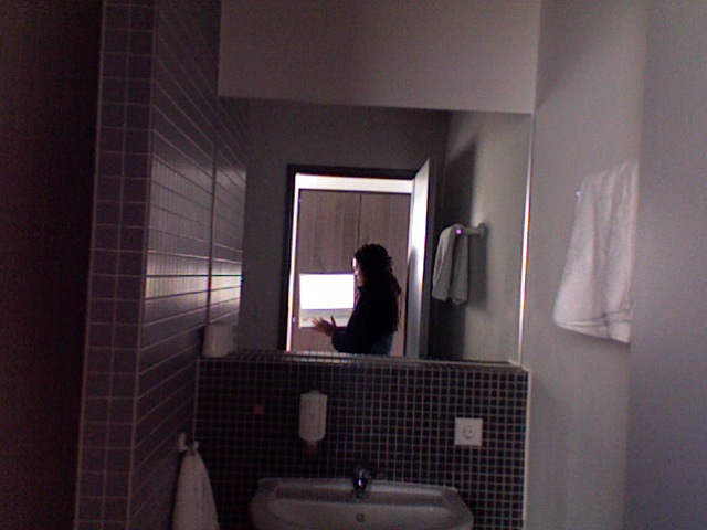 Hotel bathroom mirror-in-mirror: dark figure at the sink reflected through a doorway, grey mosaic tiles, towel on the rail Hotel bathroom mirror-in-mirror: dark figure at the sink reflected through a doorway, grey mosaic tiles, towel on the rail