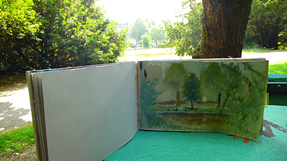 Handbound sketchbook open on a green park table: gouache painting of the pond and trees, the same real pond hazy behind Handbound sketchbook open on a green park table: gouache painting of the pond and trees, the same real pond hazy behind