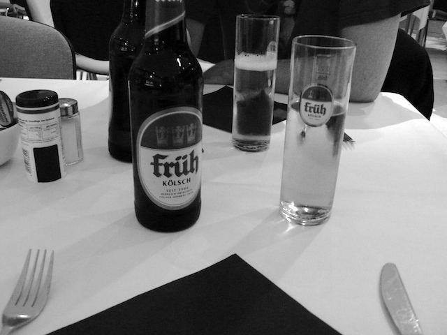 Black-and-white café table: a Früh Kölsch bottle and two tall glasses, one full, one nearly empty