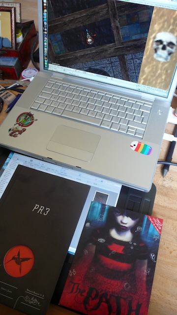 Silver laptop running a 3D level editor with skull textures; below it the boxed retail edition of The Path and a black book stamped PR3