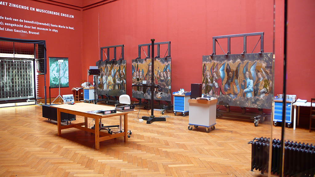 Museum conservation room, deep red walls: Memling's singing and music-making angel panels on wheeled easels among examination lamps and tool carts Museum conservation room, deep red walls: Memling's singing and music-making angel panels on wheeled easels among examination lamps and tool carts