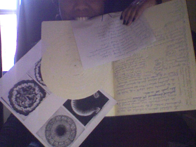 Webcam self-portrait at 18h54: a sheet of handwriting held in the teeth over an open notebook and printed mandala images