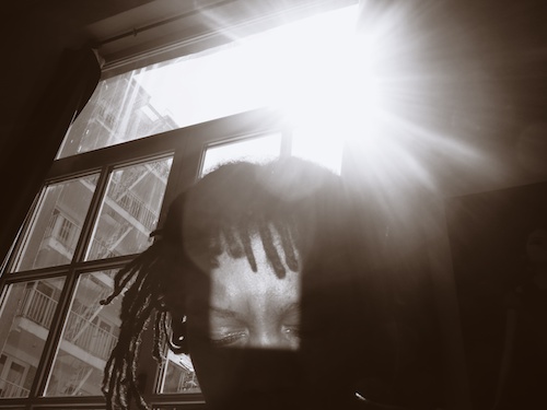 Sepia self-portrait, head bowed before a window: sunlight flares over the top of the head, dreadlocks hanging, apartment block beyond
