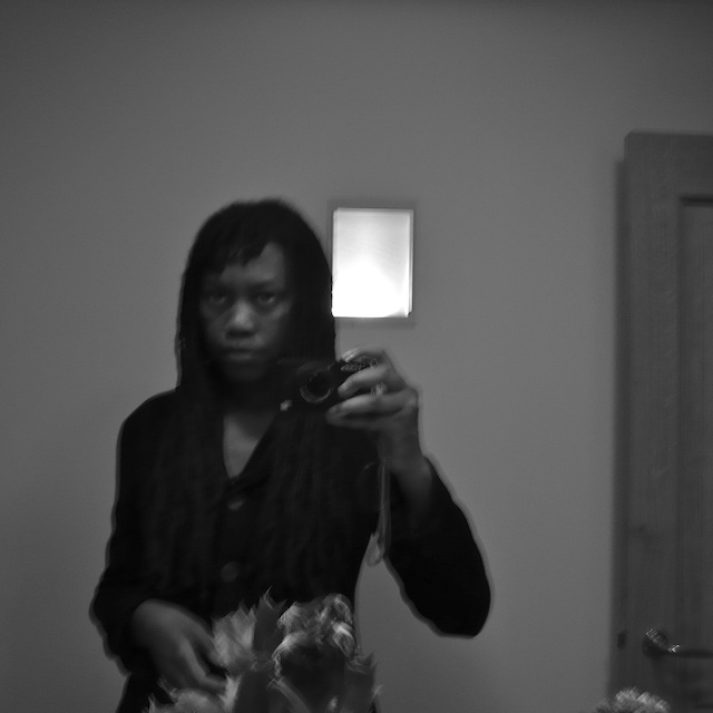 Blurred black-and-white mirror self-portrait: standing in a bare room with a camera at the chest, a lit sconce and door behind