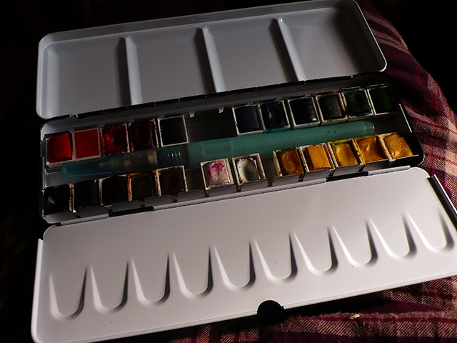 New watercolor tin open on a plaid lap: two rows of half-pans from reds through blues to ochres, a water brush laid across