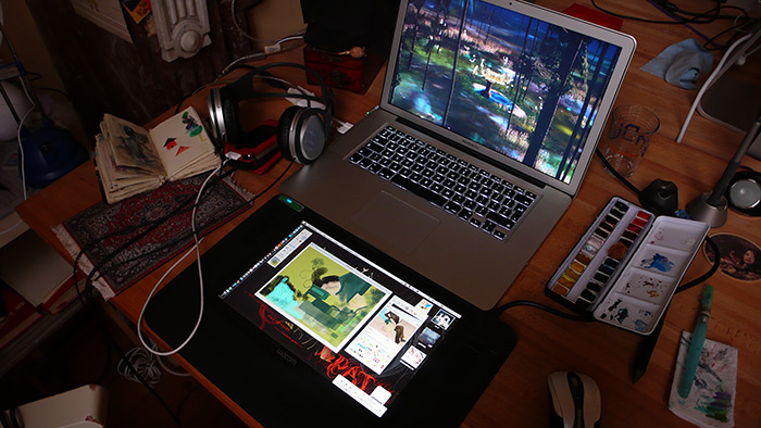 Night desk: Wacom tablet showing a painting in progress of a girl in a forest clearing, laptop with the reference scene, headphones and the watercolor tin nearby