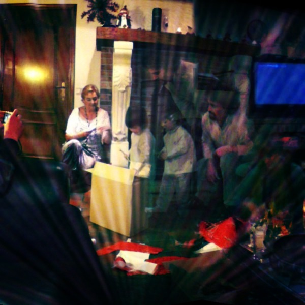 Christmas Eve double exposure: family unwrapping presents by the fireplace, a woman on the hearth, children over a big box, torn red paper on the floor Christmas Eve double exposure: family unwrapping presents by the fireplace, a woman on the hearth, children over a big box, torn red paper on the floor