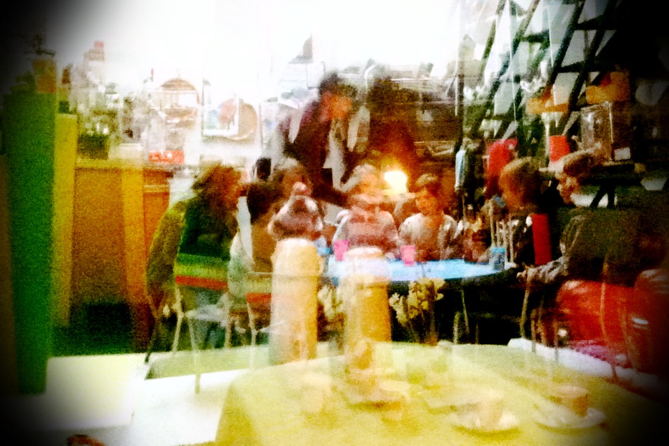 Overexposed café scene through glass: children's party around a blue table with pink cups, an adult leaning over the middle, greenhouse shelves behind Overexposed café scene through glass: children's party around a blue table with pink cups, an adult leaning over the middle, greenhouse shelves behind