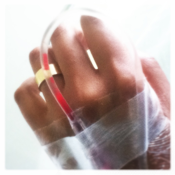 Hospital close-up: a hand taped with gauze, an IV line running red past a gold ring