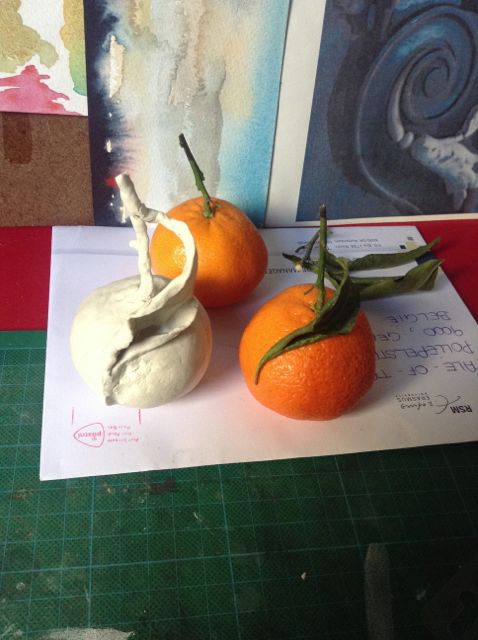 On the studio's green cutting mat: a small white clay sculpture splitting open like a fruit, posed with two clementines on an envelope