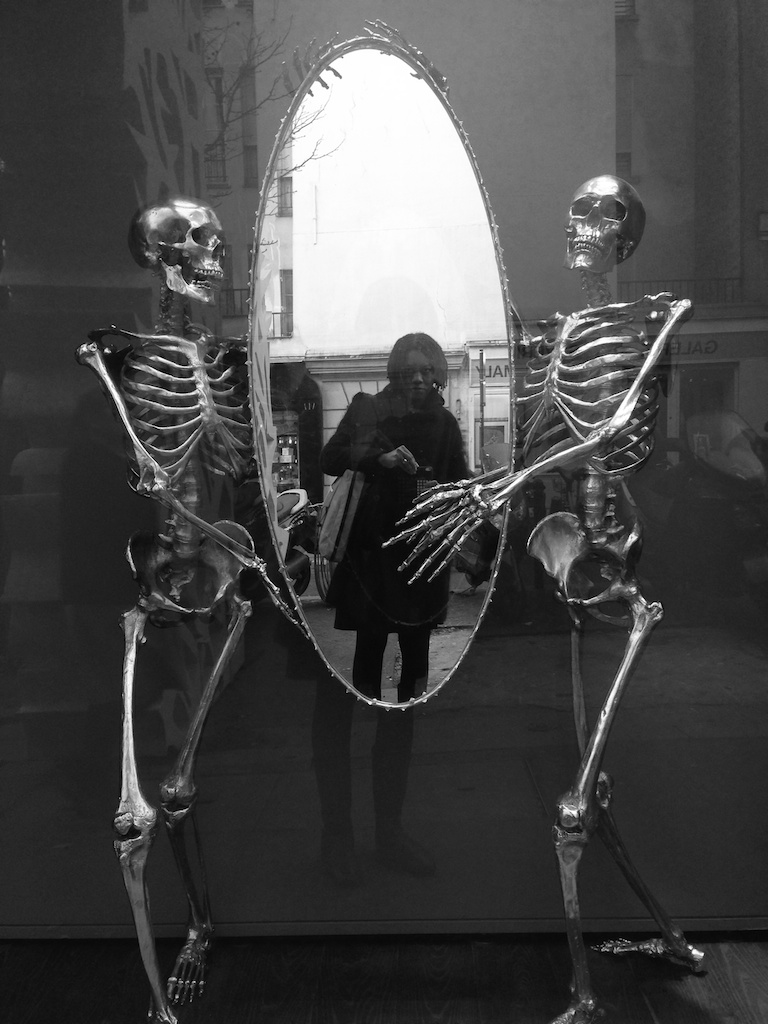 Two life-size gilded skeleton sculptures hold an oval mirror in a dark shop window; the photographer stands reflected between them, all in black