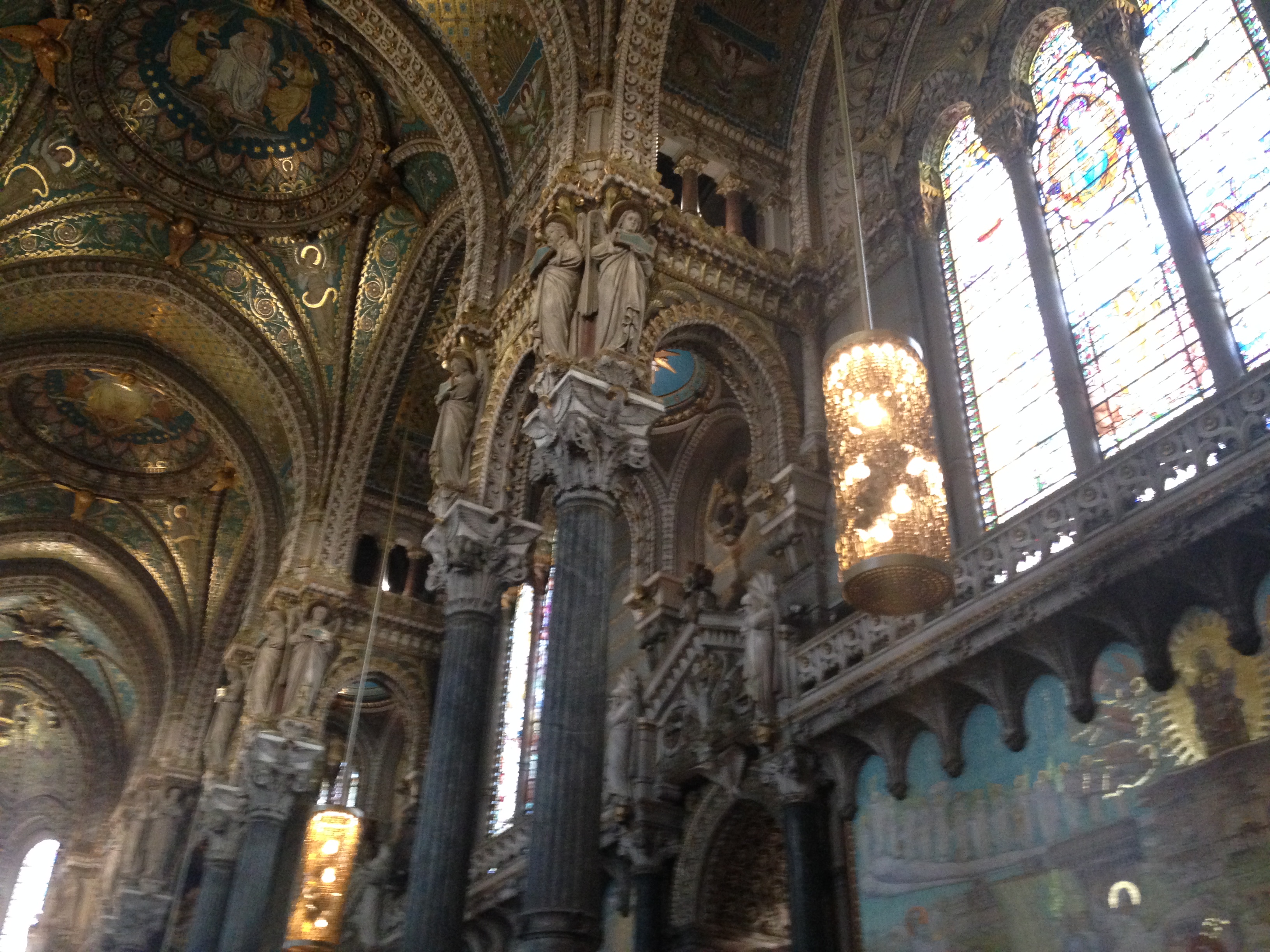 Inside the basilica of Fourvière in Lyon: blue-and-gold mosaic vaults, angel-topped columns, a glittering cylindrical chandelier against tall stained glass