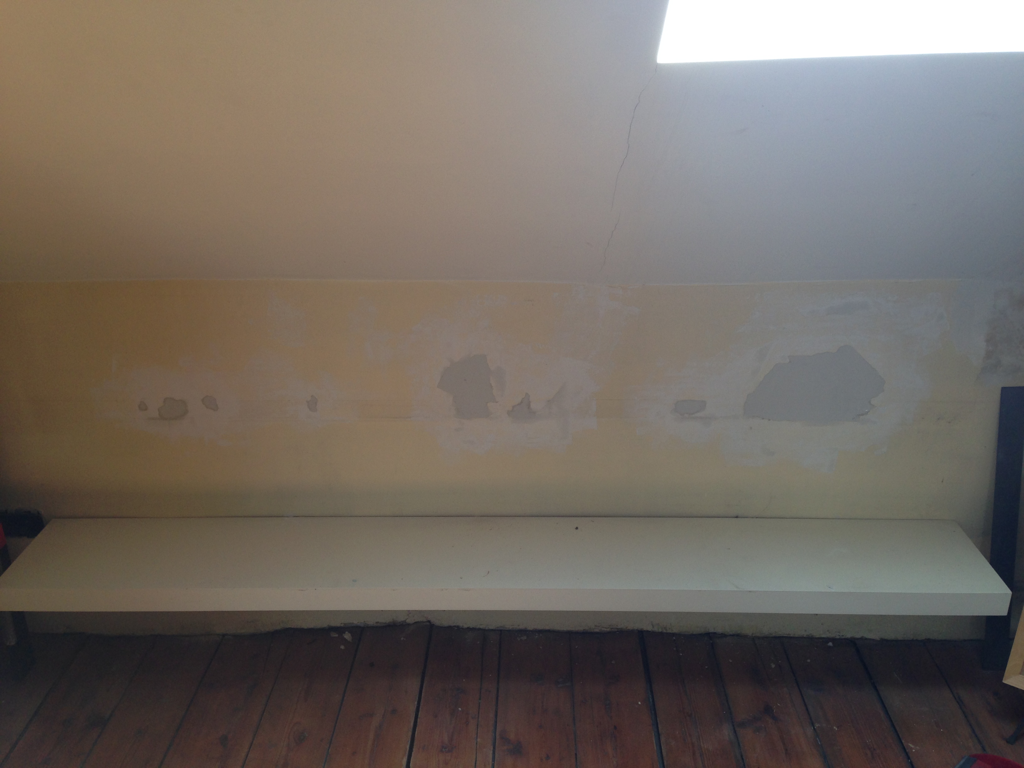 Attic wall mid-repair: patches of white filler over old yellow paint above a long white floating shelf, bare wood floor below