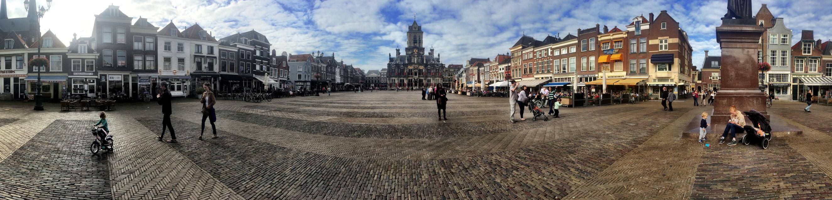 Panorama of the Markt in Delft: herringbone brick square, gabled houses all around, the town hall at center, children and strollers at the statue's plinth