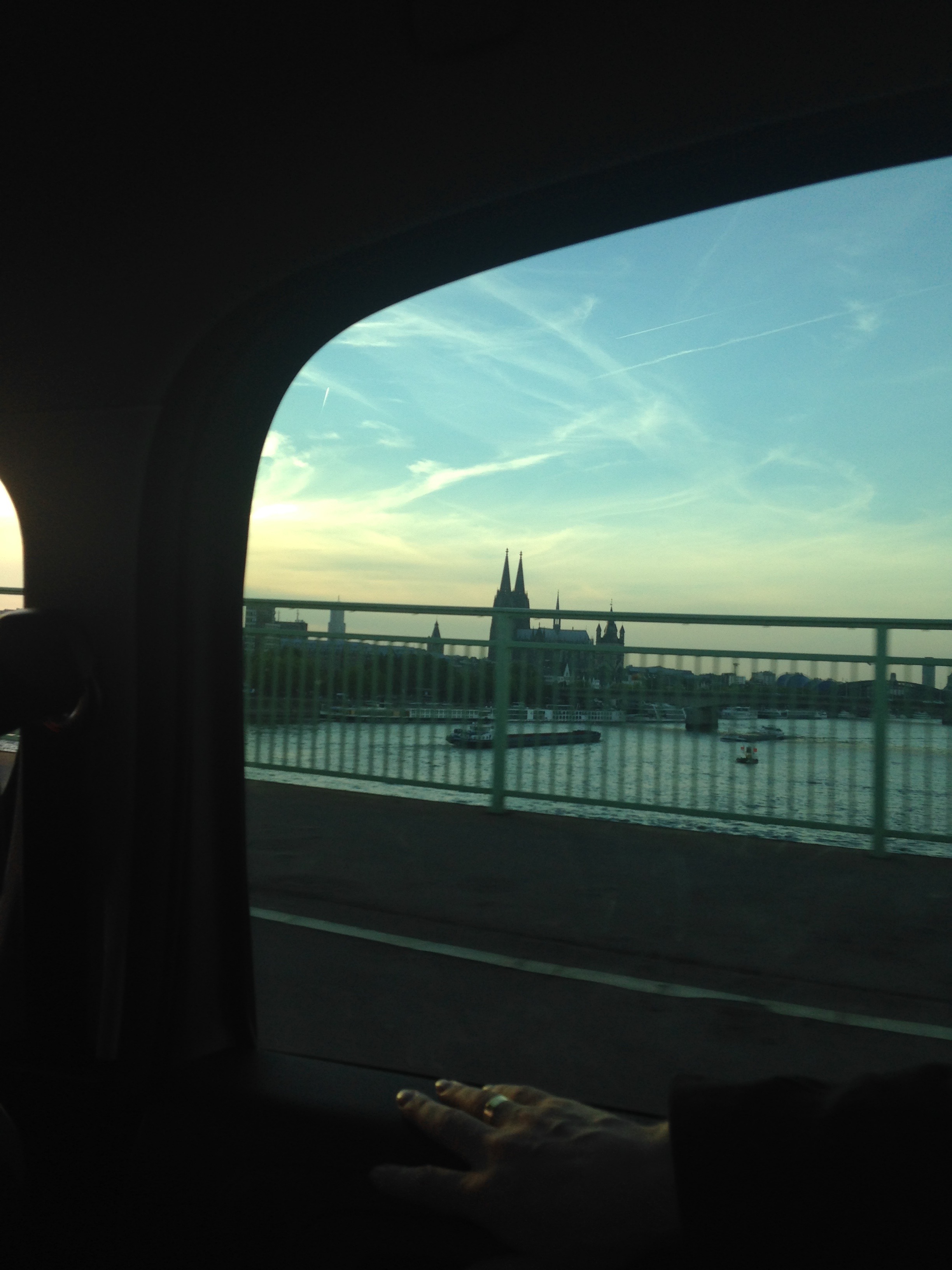 The Rhine from a car window: Cologne Cathedral's twin spires on the skyline, barges below, a hand resting on the sill in shadow