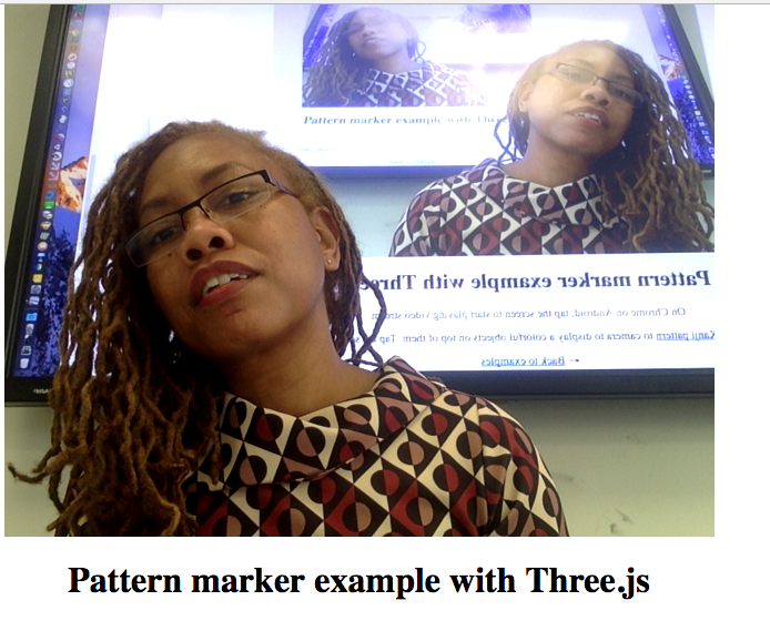 Webcam still from an AR test — 'Pattern marker example with Three.js' — peering over glasses in a patterned dress, the screen behind mirroring the page